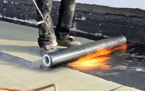 Un couvreur utilise un chalumeau pour souder un rouleau de membrane bitumineuse, créant ainsi une toiture chaude et étanche.