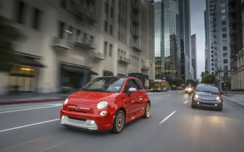 Czerwony Fiat 500e jedzie ulicą w mieście, w tle wieżowce i autobus.