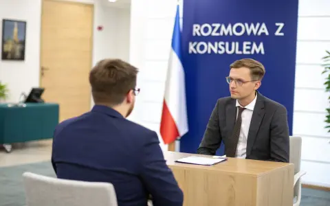 Rozmowa z konsulem. Mężczyzna w okularach i garniturze siedzi przy biurku, obok flaga.