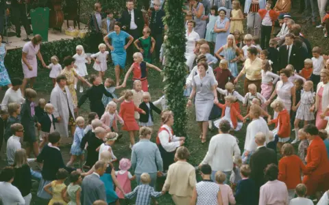 Många barn och vuxna dansar ringdans runt en midsommarstång i Sverige.