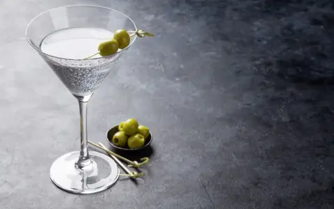 Ein Glas trockener Wermut mit Oliven garniert, daneben eine Schale mit weiteren Oliven auf dunklem Untergrund.