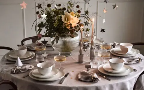 Enkel nyårsdukning med vita tallrikar, champagneglas, stjärnformade dekorationer och en blomsterbukett i en glaskaraff.