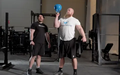 Man med skägg utför en kettlebell press med en blå kettlebell. En annan man står bredvid och tittar.