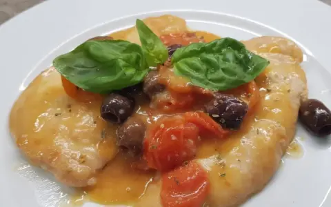 Delizioso pollo alla mediterranea con pomodorini, olive nere e basilico fresco su un piatto bianco.