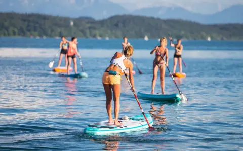 Eine Gruppe von Frauen genießt das Stand-Up-Paddling auf einem See. Sie stehen auf ihren Boards und paddeln durch das ruhige Wasser, während im Hintergrund Berge und Bäume zu sehen sind.