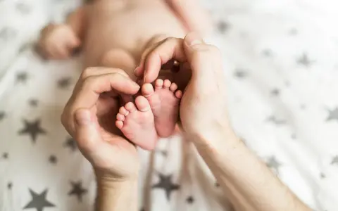 Dłonie obejmujące stópki 7 tygodniowego dziecka. Delikatne paluszki i pięty maluszka otoczone troską.