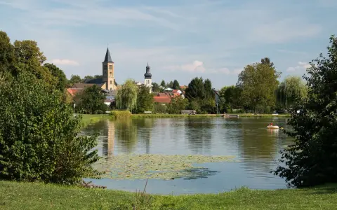 Idyllischer Elsenzer See mit Seerosen, Bäumen und einer Kirche im Hintergrund. Ein Boot mit zwei Personen treibt auf dem Wasser.