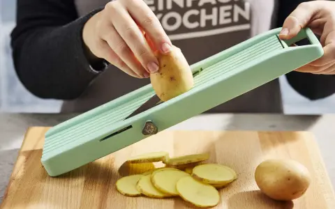 Eine Hand schneidet Kartoffeln für ein Kartoffelsandwich auf einem Hobel. Dünne Scheiben liegen auf dem Holzbrett.