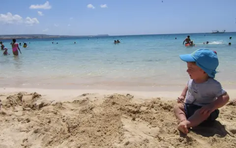 Bambino con cappellino blu gioca sulla sabbia a Cipro, mentre altri si divertono in mare.