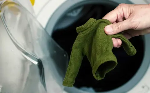 Main verte, petite, tenue dans une main, devant une machine à laver ouverte. Le coton rétrécit il au lavage ?
