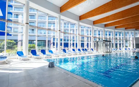 Wnętrze z basenami hotelowymi, leżakami i dużymi oknami z widokiem na budynek hotelowy.