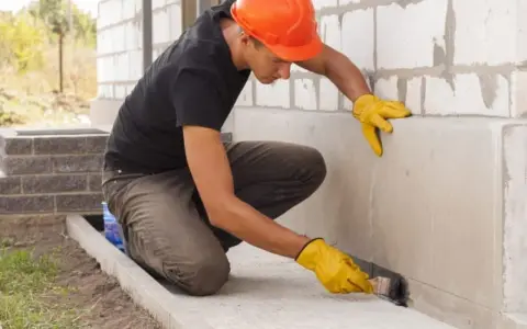 Obrero con casco naranja aplica producto para impermeabilizar pared exterior de bloque de hormigón.