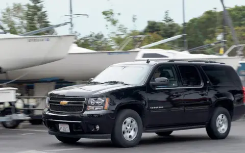 Czarny Chevrolet Suburban jedzie obok przyczep z łodziami, w tle drzewa.