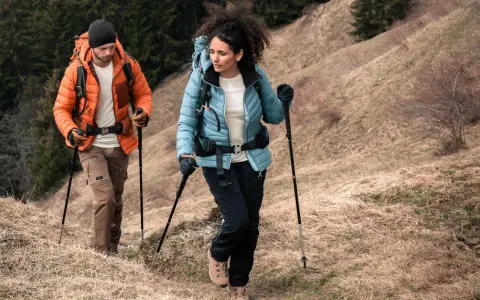 Para dobrać kijki trekkingowe do wzrostu, zwróć uwagę na kąt 90 stopni w łokciu podczas marszu. Para turystów z plecakami idzie po zboczu.