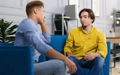 Dwóch mężczyzn siedzi na kanapie, rozmawiając. Jeden z nich opowiada o tym, jak przełamać barierę językową, szukając inspiracji w szczerej rozmowie.