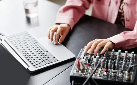 Osoba z tatuażami na dłoniach reguluje suwaki na mikserze, obok laptopa. Dowiedz się, jak podłączyć mikser do laptopa.
