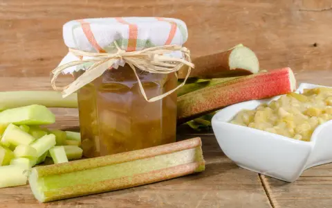 Un bocal de confiture de rhubarbe pas trop sucrée, entouré de tiges de rhubarbe fraîchement coupées et d'un bol de compote.
