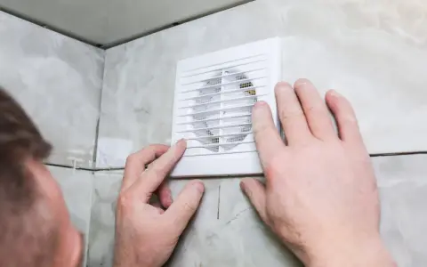 Mains d'homme ajustant une grille de VMC dans une salle de bain car une mauvaise odeur persiste.