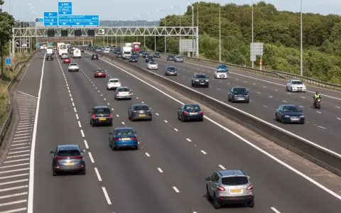 Autostrada z ruchem lewostronnym, pełna samochodów i ciężarówek. Znaki drogowe wskazują M1, A405 i M25.