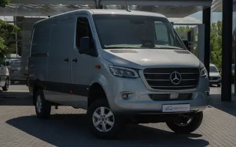 Srebrna budowa ciężarówki, model Mercedesa Sprintera, stoi na parkingu przed nowoczesnym budynkiem.