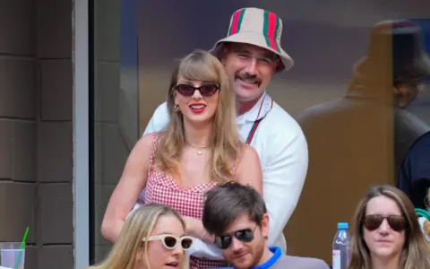 Travis Kelce e Taylor Swift sorridono, lei con un vestito a quadri rossi, lui con un cappello Gucci.