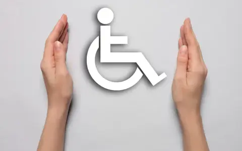 Dwie dłonie otaczają biały symbol osoby na wózku inwalidzkim, symbolizując wsparcie i akceptację dla osób z niepełnosprawnościami.