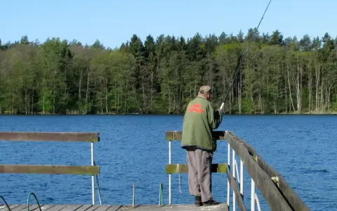 Wędkarz na pomoście nad spokojnym zalewem Koronowskim, w tle las.