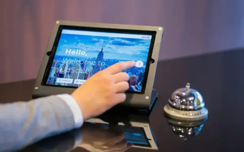 Dłoń dotyka ekranu tabletu z napisem "Welcome to UpScale". Czy można zarezerwować hotel w wieku 17 lat? Obok dzwonek hotelowy.