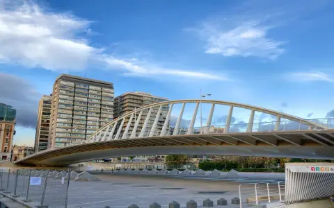 El moderno puente de la Exposición cruza un río, con edificios altos al fondo bajo un cielo azul con nubes.