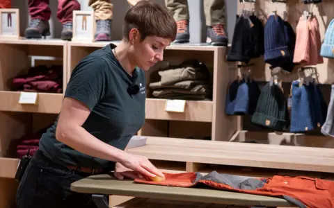 Mitarbeiterin pflegt orangefarbene Hose, damit sie für den Verkauf wie neu wachsen kann. Im Hintergrund sind Kleidung und Mützen ausgestellt.