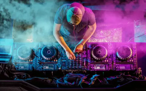 DJ con cuffie e cappellino mixa musica hause su console illuminata da luci viola e blu, circondato da fumo.