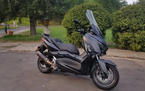 Czarny skuter Yamaha XMAX 300 zaparkowany na tle zieleni i drzew.
