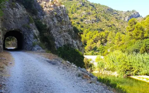 Camino de grava junto a un río y un túnel en la vía verde de Alicante. Naturaleza exuberante y rocas escarpadas.