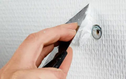En hand använder en spackel för att spackla hål i en vit vägg.