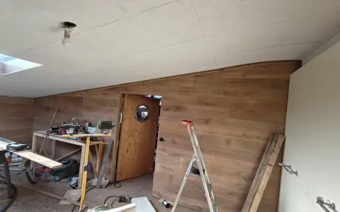Taket är målat OSB. Rummet är under renovering med träpanel på väggarna, en dörr med runt fönster och byggmaterial utspritt.