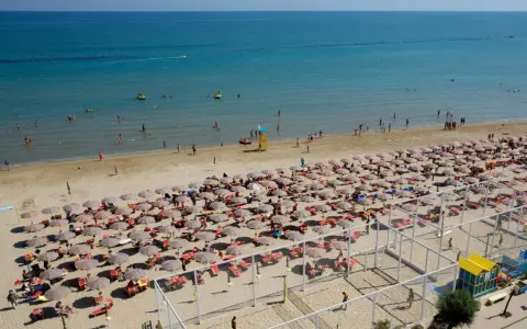 A crowded beach with rows of umbrellas and sunbathers, and people swimming in the blue sea.