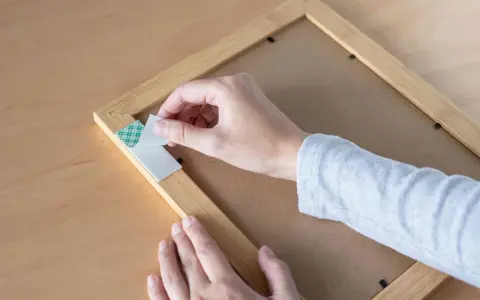 Mano retirando cinta de doble cara de un marco de madera. Aprende cómo quitar cinta de doble cara de la pared sin dañarla.