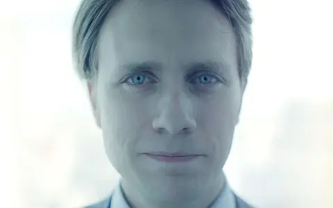 Mężczyzna o niebieskich oczach, z lekko uśmiechniętymi ustami, wygląda jakby był pracownikiem evil corp.