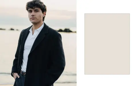 Davide Avolio, con le mani in tasca, indossa un cappotto scuro e una polo bianca, con il mare sullo sfondo.