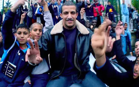 Un chanteur rap français, entouré de jeunes fans, pose avec confiance.
