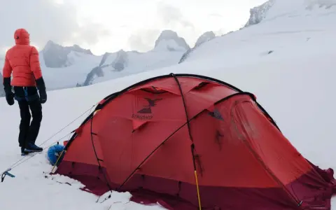 Alpinista w czerwonej kurtce stoi obok namiotu na tle ośnieżonych szczytów. W oddali widać groźne góry, gdzie mogą wystąpić lawiny w Alpach.