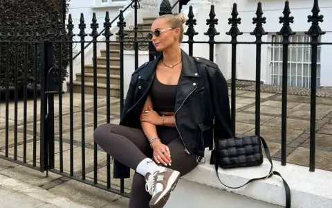 Kobieta w skórzanej kurtce i legginsach, uosobienie stylu athleisure, siedzi na murku przed kamienicą.
