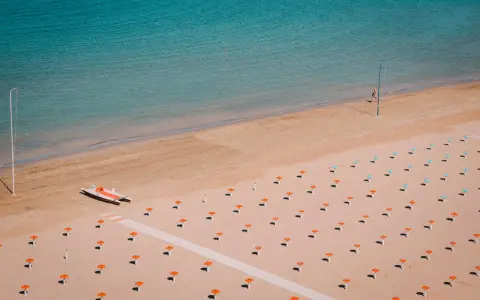 Idealne loty do Rimini: pusta plaża z rzędami parasoli, łódka na piasku i turkusowe morze.