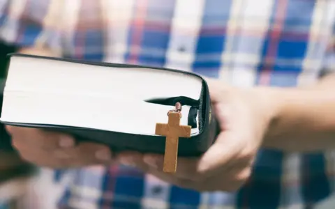 Osoba w kratę trzyma Biblię z drewnianym krzyżem, symbolizującym dwa przykazania miłości.