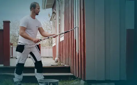 Man i arbetskläder använder en lång roller för att måla huset. En hink med färg står på gräset.