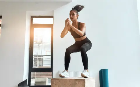 Kobieta w stroju sportowym wykonuje przysiad na skrzyni treningowej w jasnym pomieszczeniu.