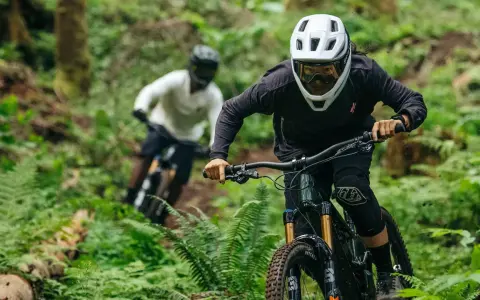 Riders in helmets and protective gear shredding a forest trail. This image shows what to wear mountain biking: full-face helmets, goggles, long sleeves, and knee pads.