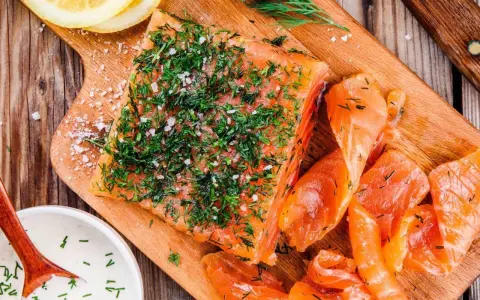 En bit gravad lax, generöst toppad med dill och flingsalt, ligger på en träskärbräda bredvid skivor av lax och en skål med sås.