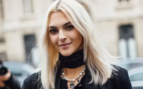 Kobieta z platynowym blondem, w czarnym golfie i naszyjnikiem z pereł, uśmiecha się lekko.