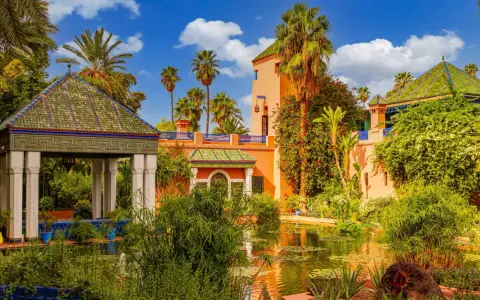 Jardin luxuriant avec un étang reflétant une maison bleue à Marrakech, palmiers et ciel bleu.